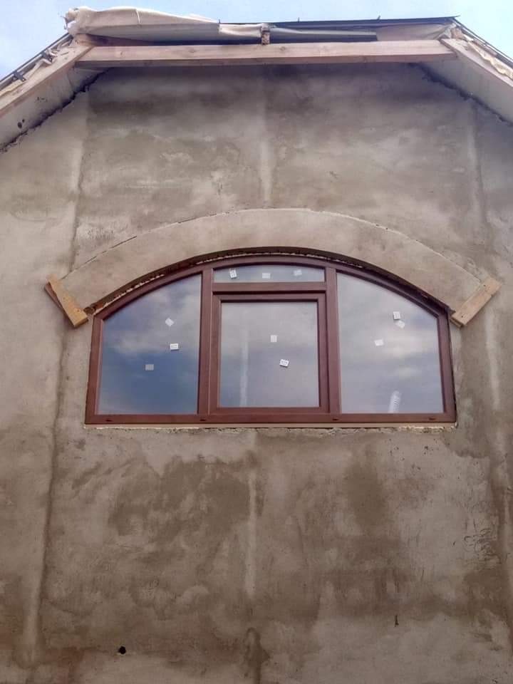 Nowe, brązowe okno o łukowym kształcie zamontowane w surowej, otynkowanej ścianie domu w trakcie budowy, widoczne drewniane elementy konstrukcyjne dachu.