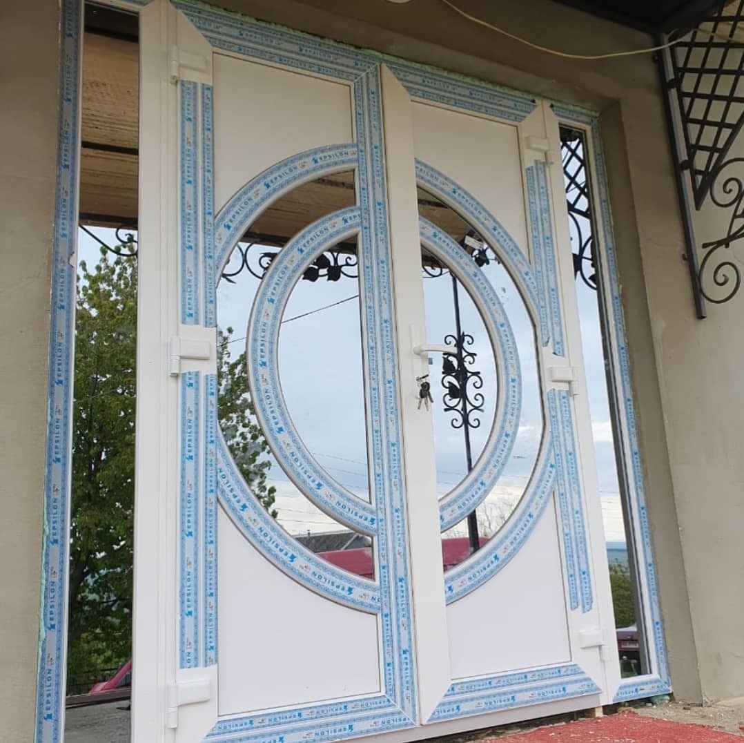 Nowe, białe drzwi wejściowe z PCV z okrągłymi, szklanymi wstawkami, widoczne klucze w zamku, niebieskie detale wykończeniowe, zewnętrzny widok.