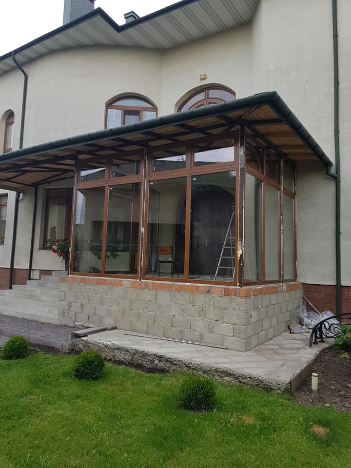 Wznoszona weranda z oknami PCV o brązowych ramach, widoczna drabina wewnątrz konstrukcji, surowy stan wykończenia elewacji i podłoża.