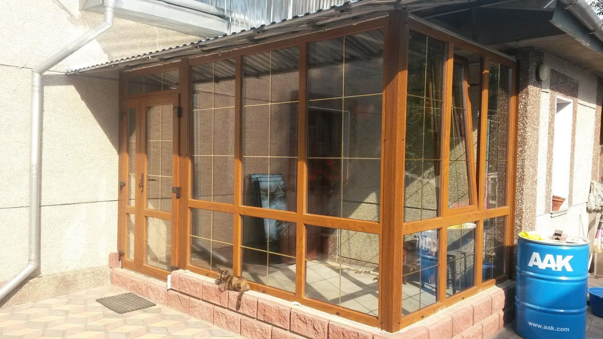 Zadaszony taras z ramami okiennymi w kolorze drewna, z widocznym kotem leżącym na murku i niebieską beczką z logo firmy w tle.