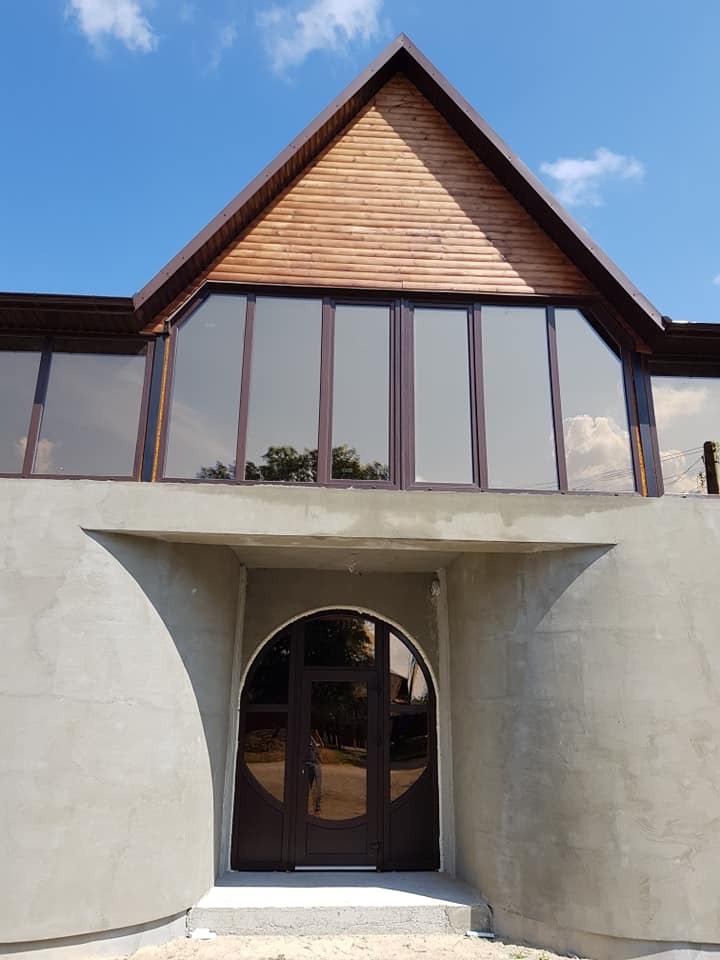 Fasada budynku w trakcie wykończenia z dużymi oknami z brązowymi ramami i okrągłymi drzwiami wejściowymi, widoczne surowe ściany z betonu.