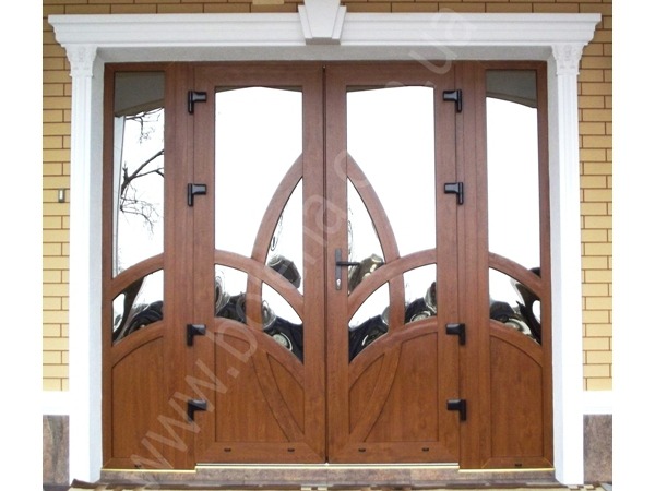 Dwuskrzydłowe, brązowe drzwi z przeszkleniami o dekoracyjnym, łukowym wzorze, osadzone w białej ramie z dekoracyjnymi kolumnami, widoczne z zewnątrz na tle jasnej cegły.