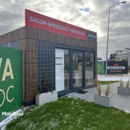 Przenośny pawilon sprzedaży mieszkań Ronson Development, z elewacją imitującą drewno, siatką na elewacji i szyldem reklamowym, otoczony trawnikiem i chodnikiem z kostki brukowej.