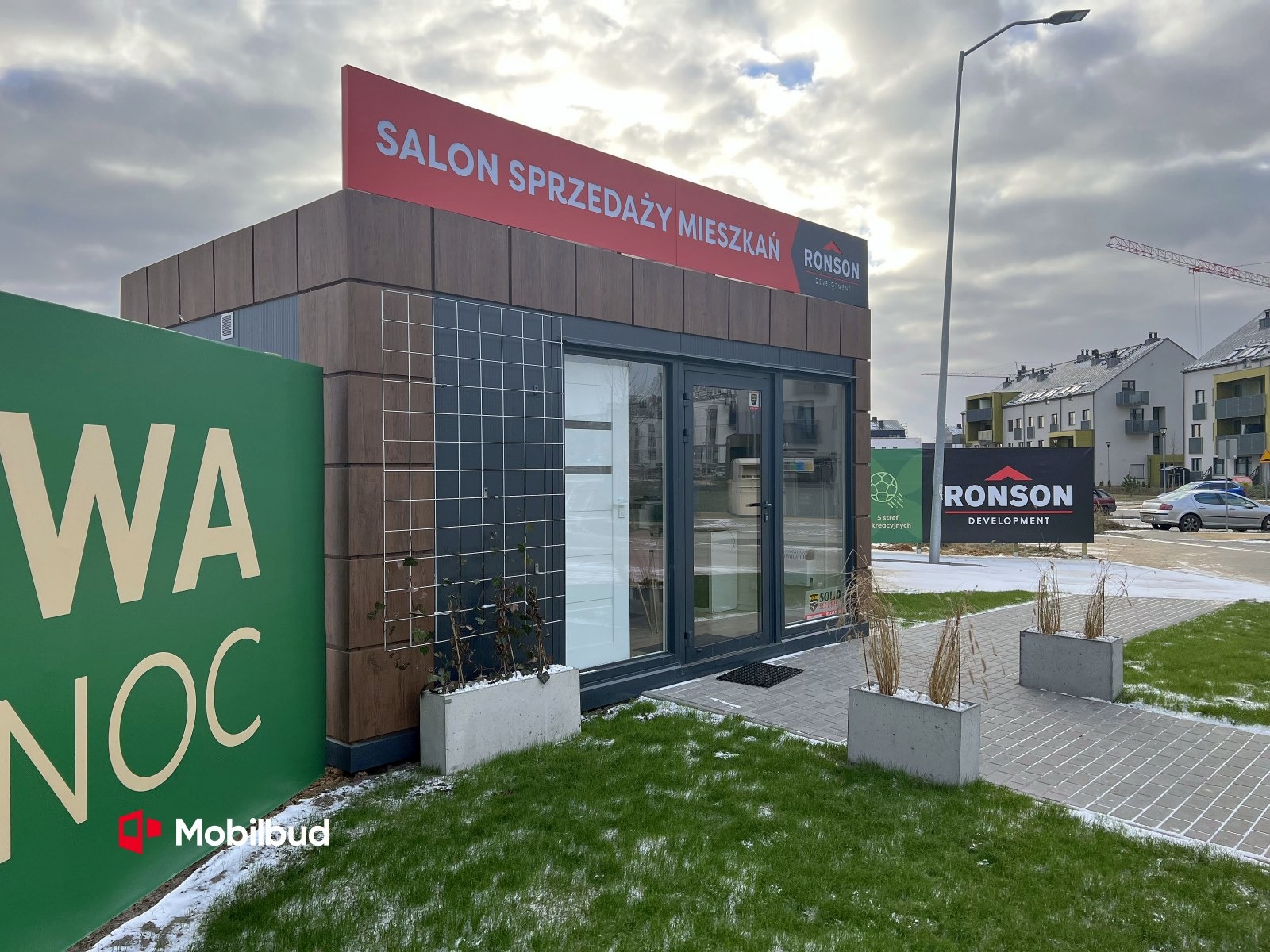Przenośny pawilon sprzedaży mieszkań Ronson Development, z elewacją imitującą drewno, siatką na elewacji i szyldem reklamowym, otoczony trawnikiem i chodnikiem z kostki brukowej.