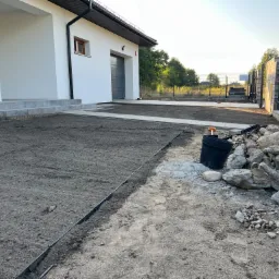 Przygotowanie podłoża pod trawnik przed nowoczesnym domem z białymi ścianami, widoczne obrzeża trawnikowe i hałda kamieni.