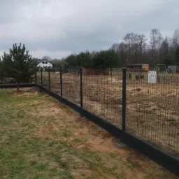 Grafitowe panele ogrodzeniowe z podmurówką i słupkami, widok na posesję z domem i drzewami w tle, pochmurny dzień.