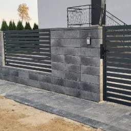 Nowoczesne ogrodzenie posesji z szarej kostki i metalowych, poziomych paneli, brama wjazdowa z domofonem, widok z zewnątrz.