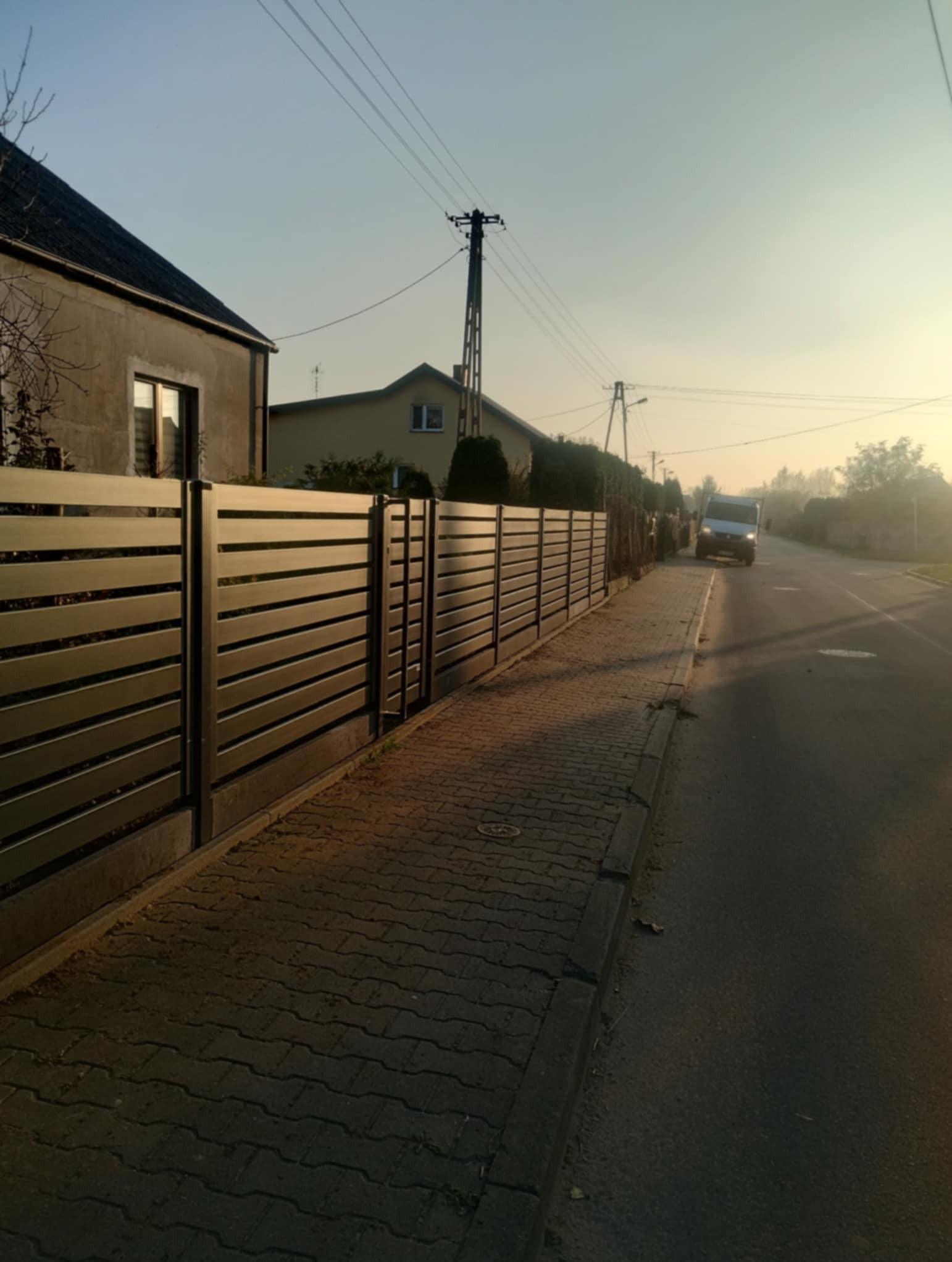 W blasku słońca. Ogrodzenie palisadowe. Na dole płyta betonowa mocowana na łączniku metalowym.