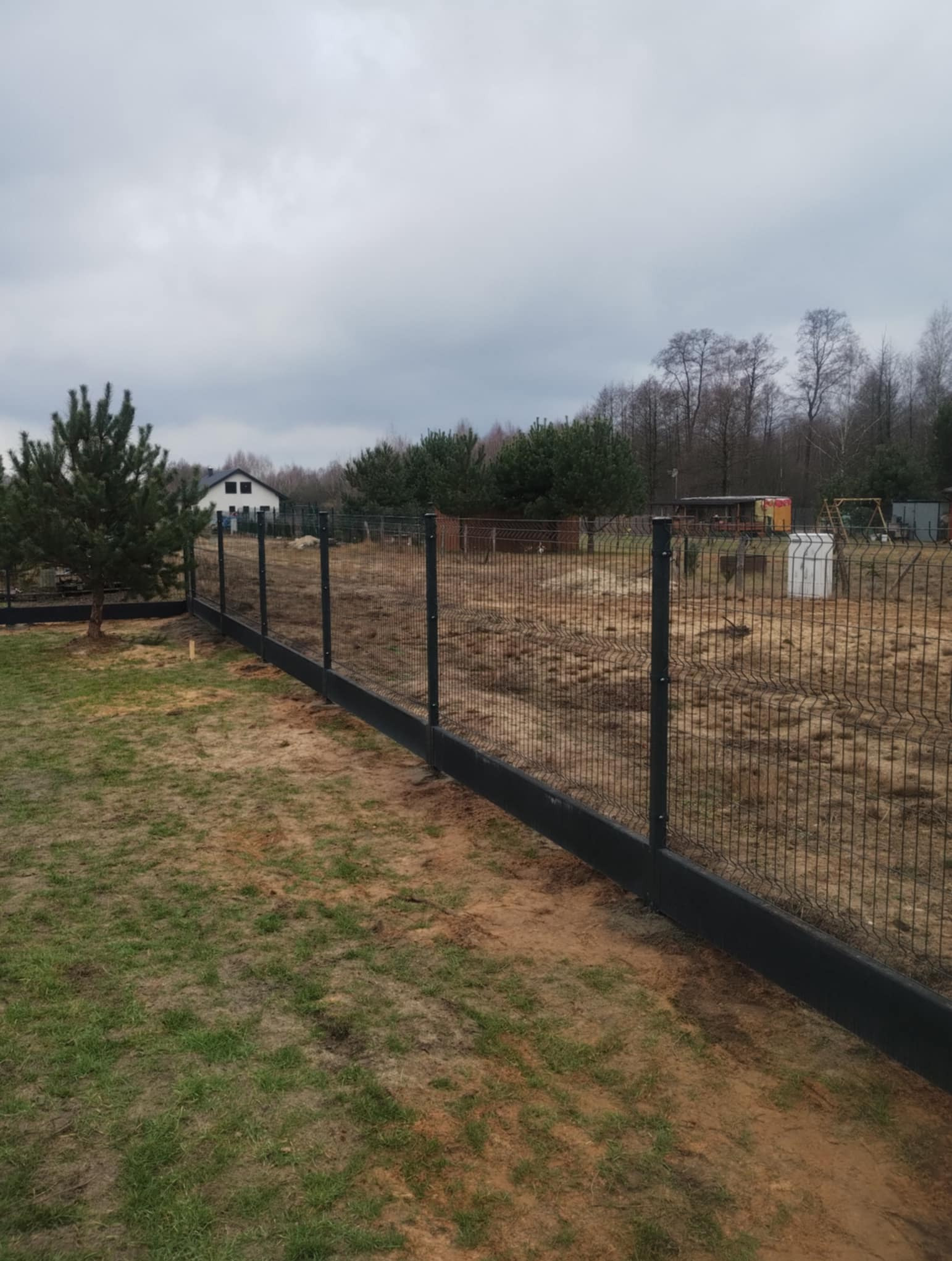 Grafitowe panele ogrodzeniowe z podmurówką i słupkami, widok na posesję z domem i drzewami w tle, pochmurny dzień.