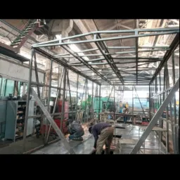 Spawanie metalowej konstrukcji szkieletowej w hali przemysłowej, iskrzenie podczas cięcia, widoczne elementy konstrukcyjne i inne osoby w tle.