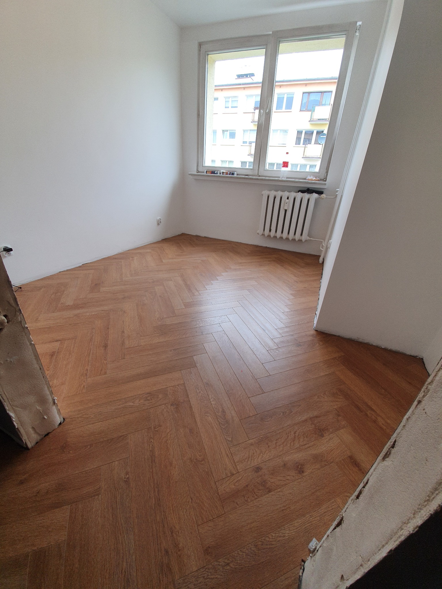 Parkiet ułożony we wzór jodełki w jasnym pokoju z grzejnikiem pod oknem, widok z perspektywy narożnika pomieszczenia.