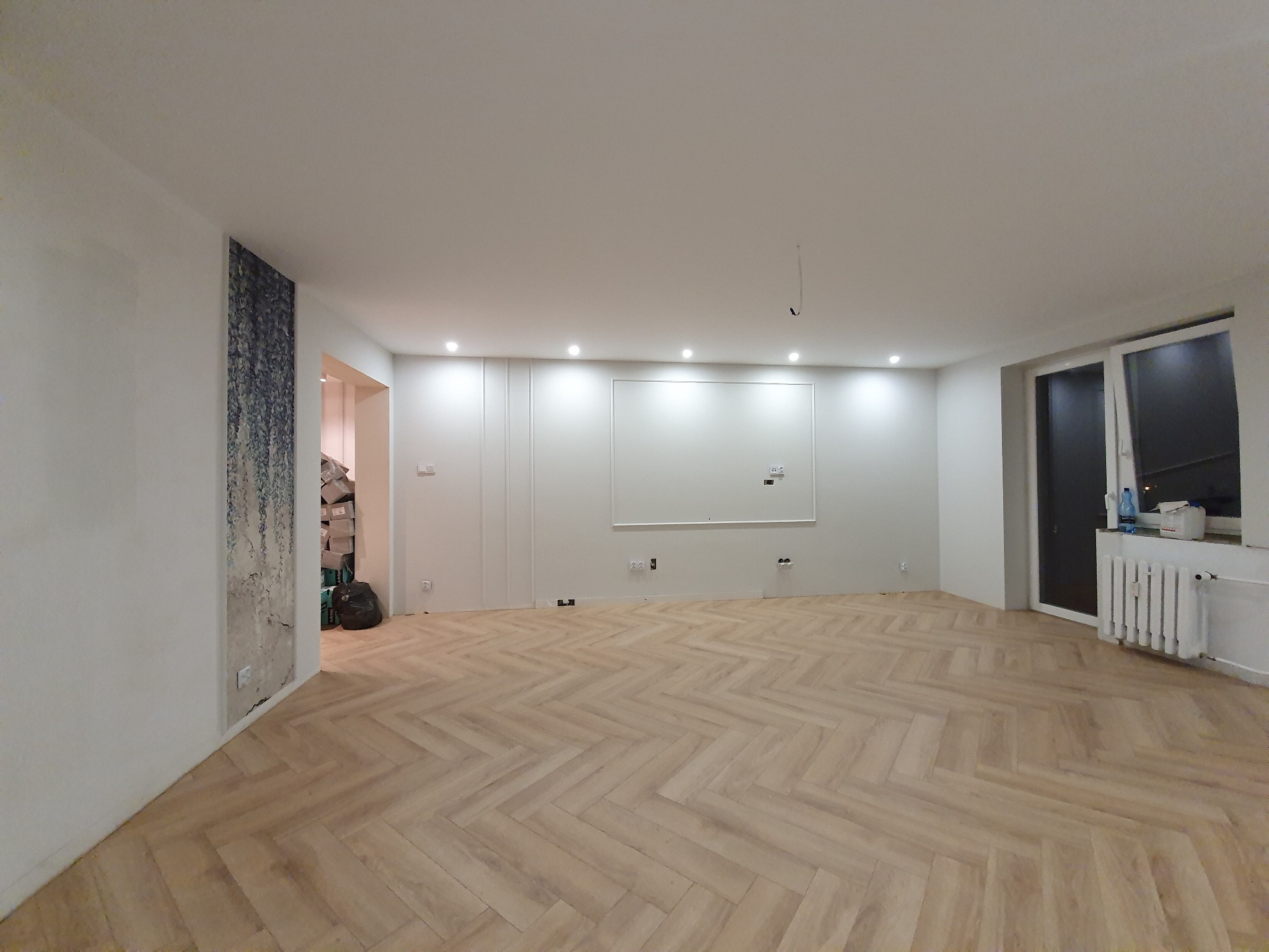 Jasny parkiet ułożony we wzór jodełki w przestronnym, jasnym pokoju z białymi ścianami i dekoracyjną tapetą, widoczne gniazdka elektryczne na ścianach.