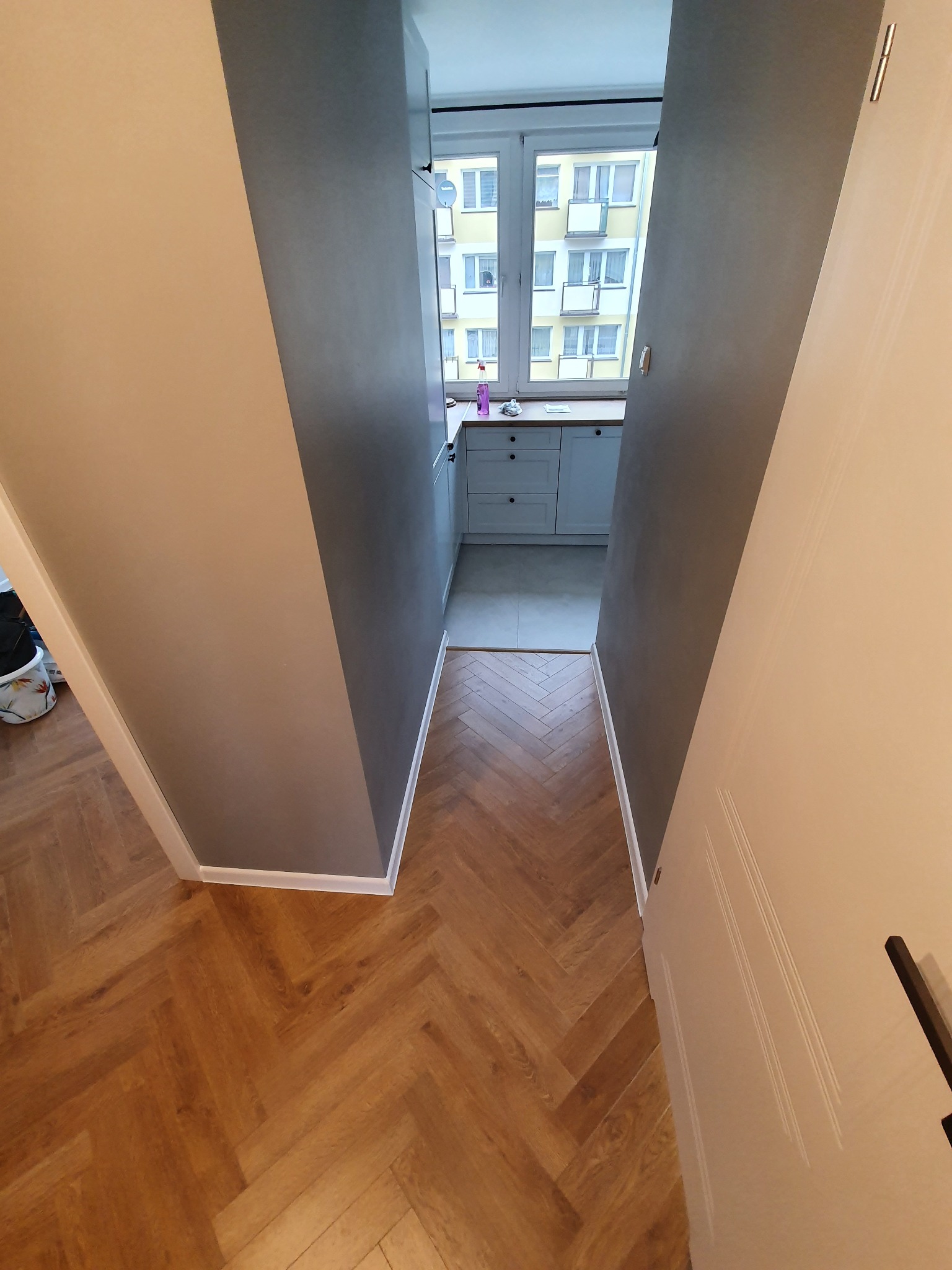 Ułożona pod kątem jodełki podłoga z paneli laminowanych w korytarzu, widok na kuchnię z białymi szafkami i oknem na blok.