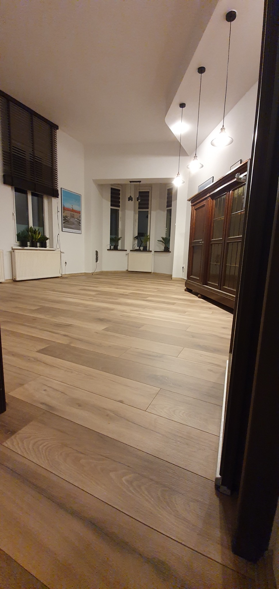 Jasne panele podłogowe ułożone w przestronnym salonie z ciemnymi drewnianymi meblami i trzema oknami z żaluzjami.