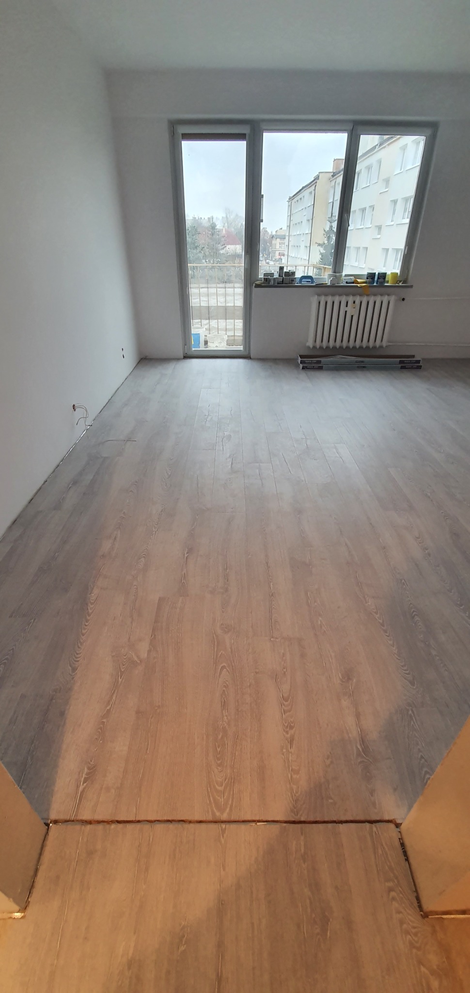 Jasna, pusta przestrzeń z nowo położoną podłogą imitującą drewno, widok na okno balkonowe z budynkami w tle, grzejnik pod oknem.