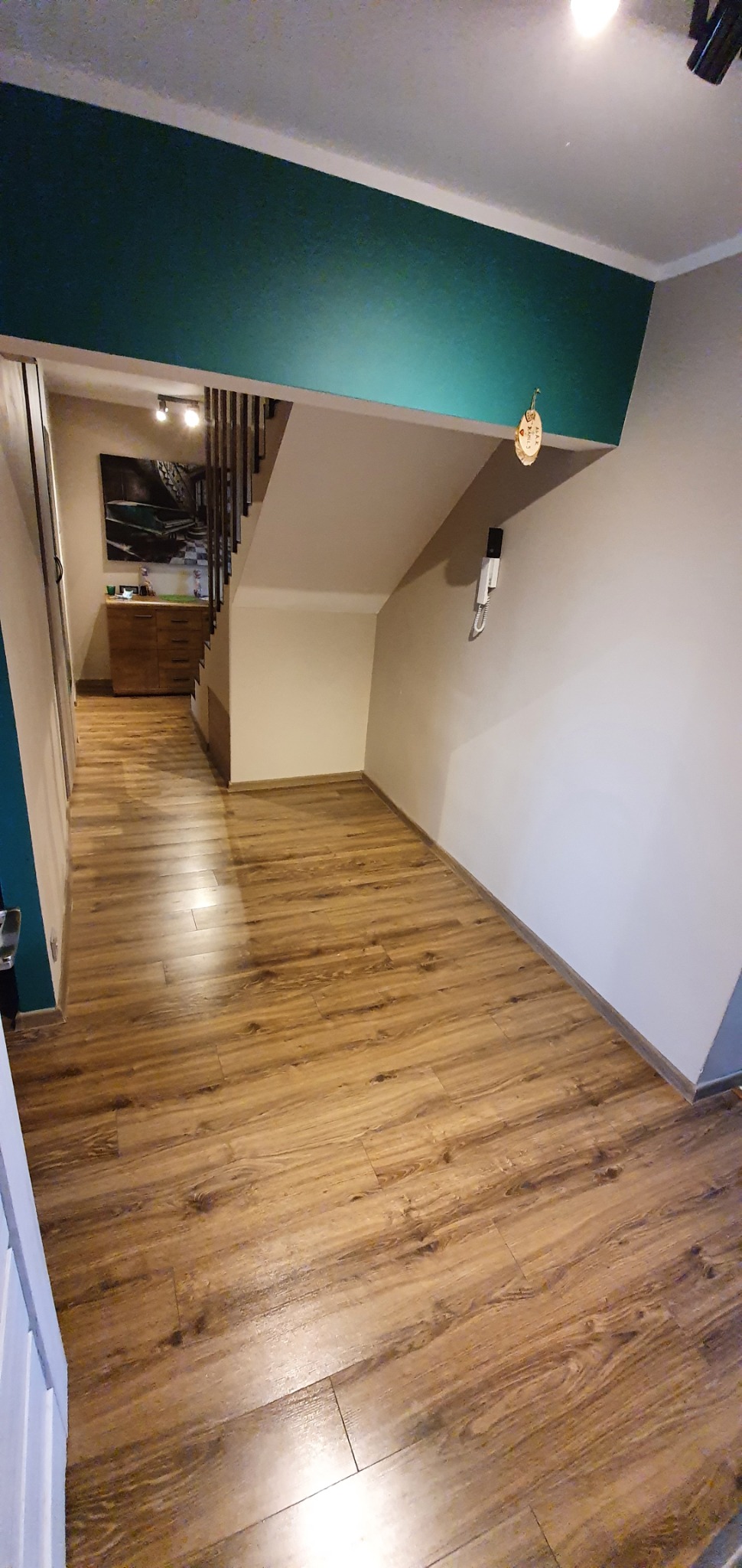 Ukończona podłoga z paneli laminowanych w odcieniach brązu, widok na korytarz z drewnianymi schodami i zieloną ścianą akcentującą.