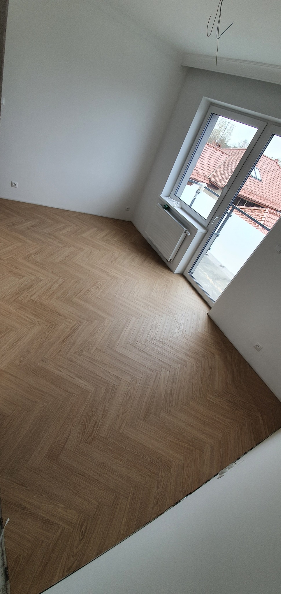 Jasne, drewnopodobne panele ułożone we wzór jodełki w pokoju z białymi ścianami i oknem z widokiem na czerwony dach.