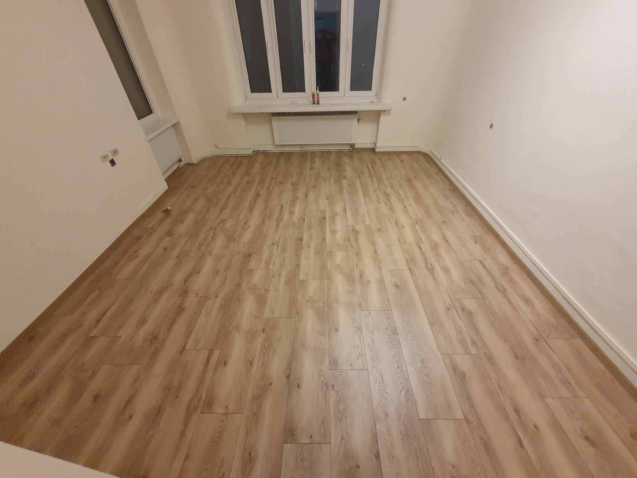 Jasne panele podłogowe w pustym, odnowionym pokoju z białymi ścianami i oknem z grzejnikiem.