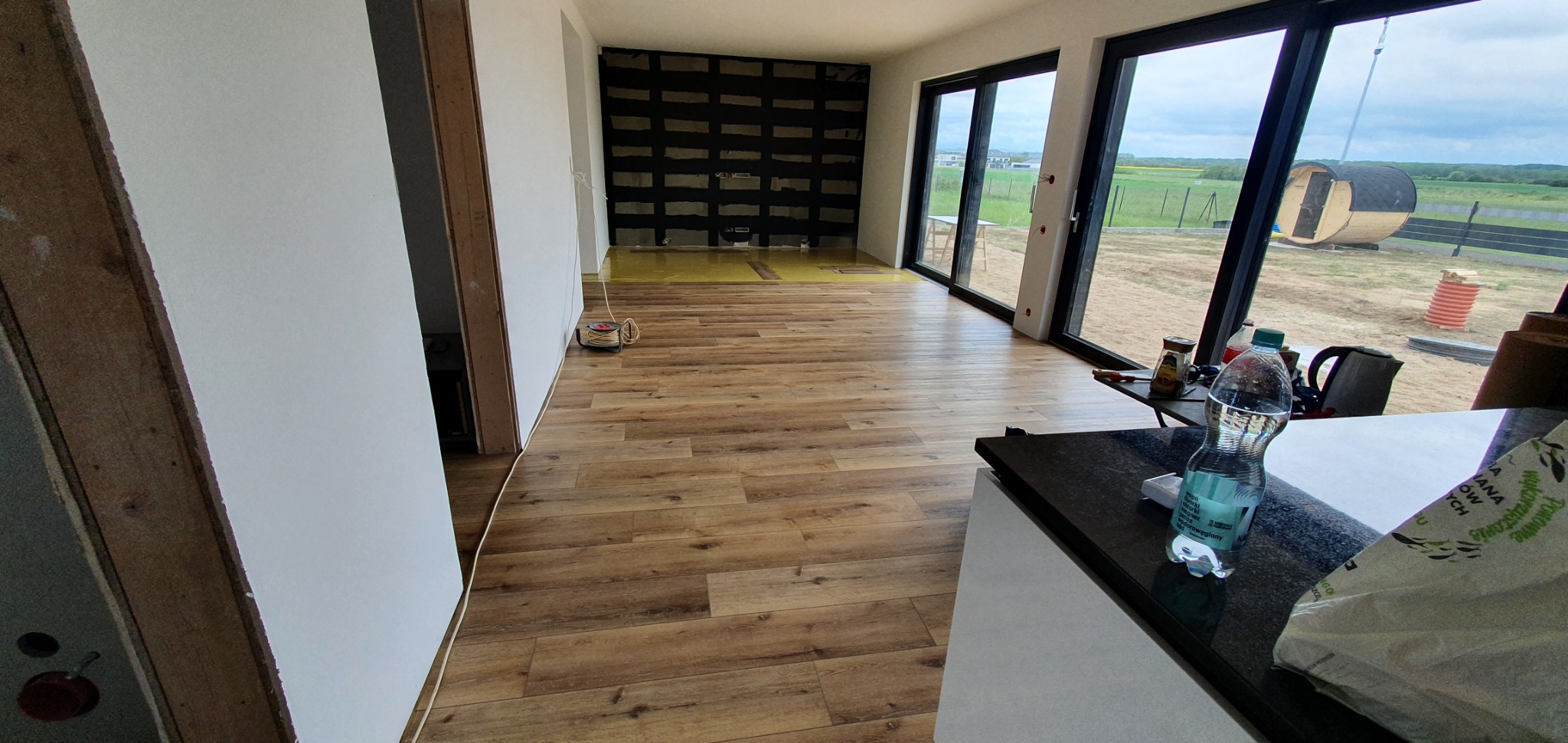 Układanie paneli podłogowych w jasnym wnętrzu domu z widokiem na saunę beczkę na zewnątrz, widoczna rolka kabla elektrycznego i butelka wody na blacie.