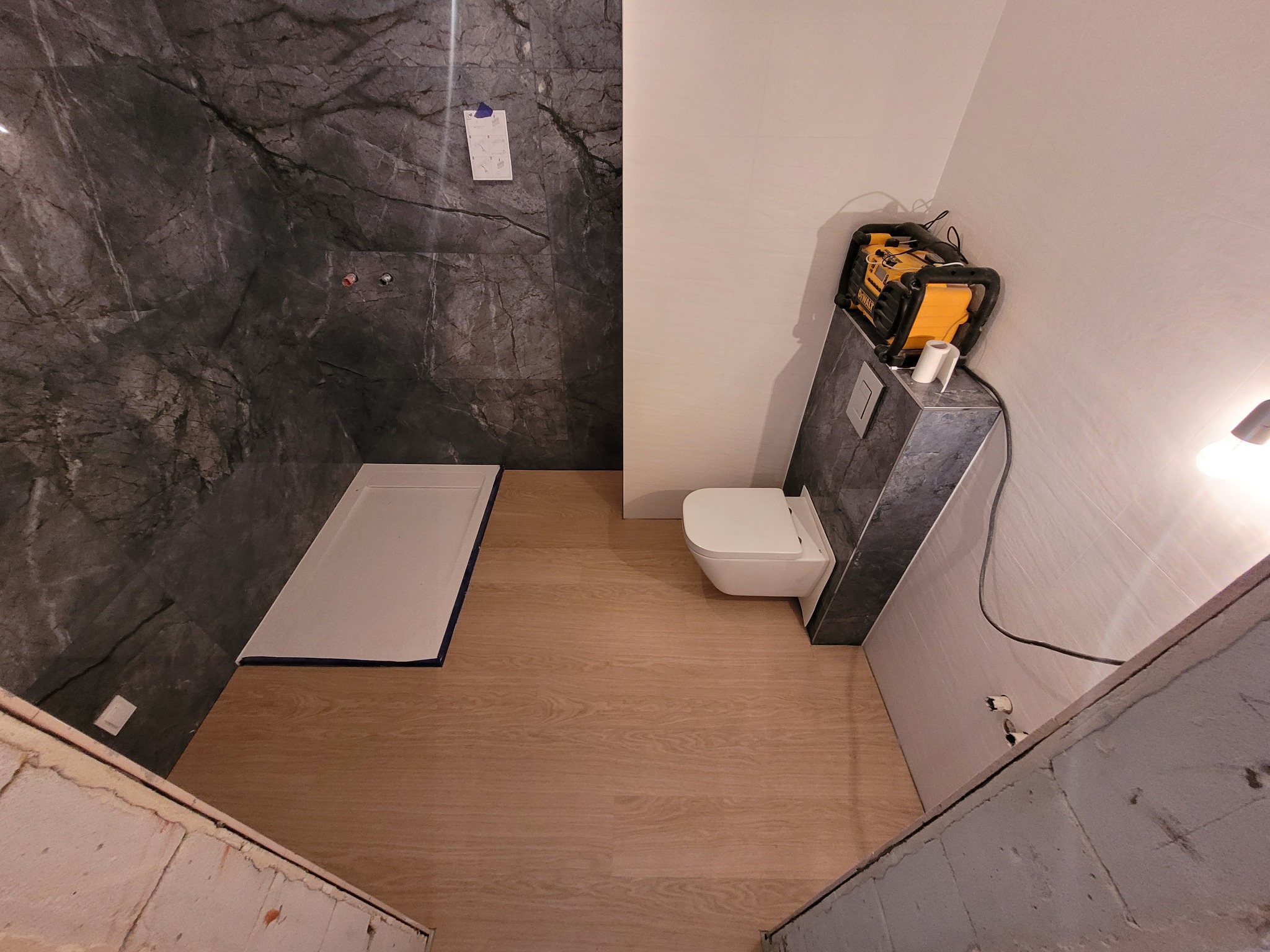 Łazienka w trakcie remontu: ułożona podłoga z paneli, zamontowany brodzik i toaleta, ściany wyłożone płytkami imitującymi marmur, widoczny laser krzyżowy DeWalt.