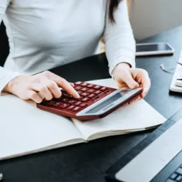 Osoba w białej bluzce używa kalkulatora w kolorze burgundowym nad otwartym notesem w otoczeniu biurowym z laptopem i stosem notatników.