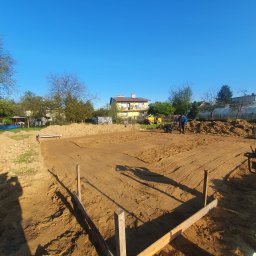 Coreltb Builders Sp. z o.o. - Przygotowanie terenu pod fundamenty: wyrównany piasek z widocznymi śladami ubijarki w kształcie przyszłego budynku, drewniane szalunki, ubijarka wibracyjna obok, pracownik w oddali i dom w tle...