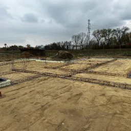 Zbrojenie fundamentów domu jednorodzinnego, widoczne stalowe pręty tworzące szkielet, szalunki z białego styropianu, przygotowany teren pod budowę w pochmurny dzień.