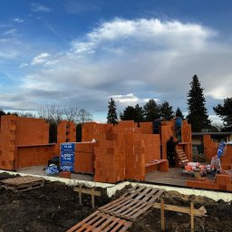 Coreltb Builders Sp. z o.o. - Wznoszenie ścian parteru domu z czerwonej cegły. Widoczne palety z cegłami Leier Plan, trzech robotników na rusztowaniu i fragmenty konstrukcji. Pochmurne niebo w tle.