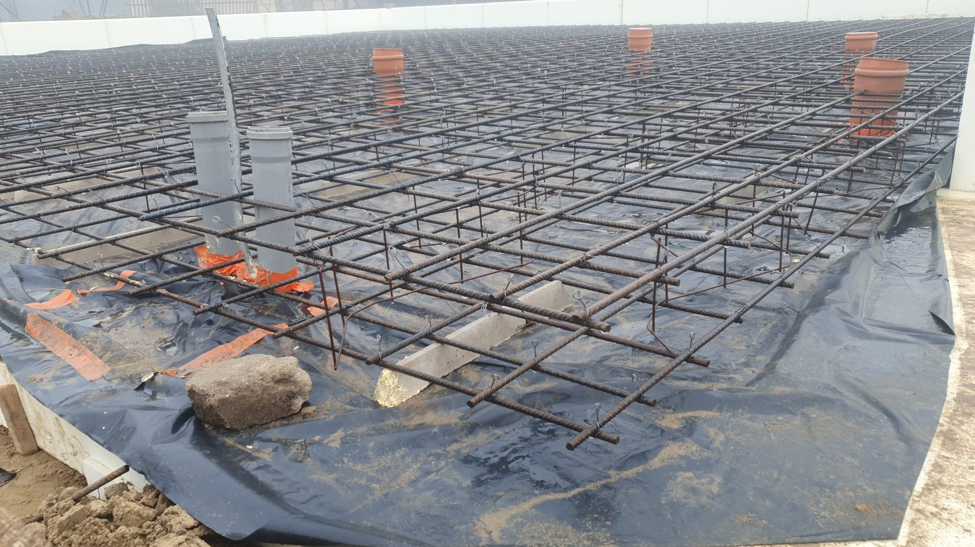 Zbrojenie fundamentu z wystającymi rurami kanalizacyjnymi, zabezpieczone folią hydroizolacyjną i betonowymi podkładkami dystansowymi.