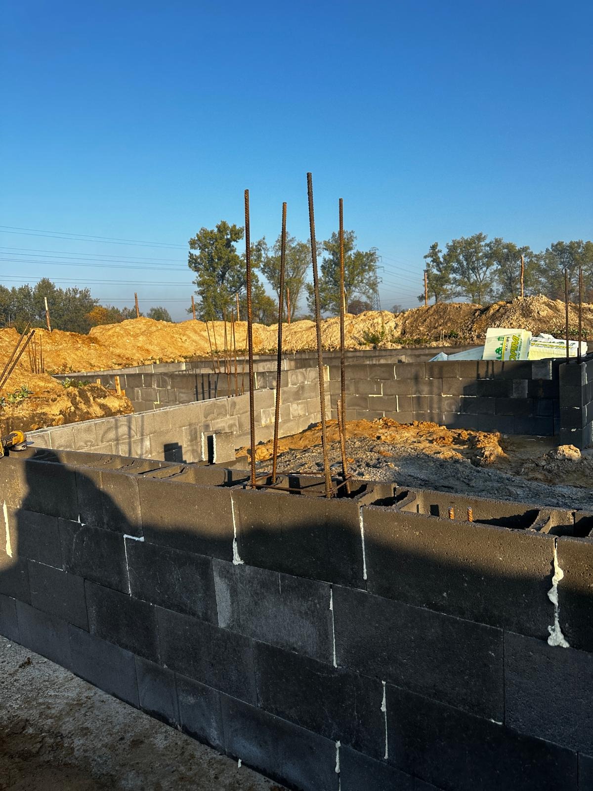 Wznoszone ściany fundamentowe z szarych bloczków betonowych, wzmocnione pionowymi prętami zbrojeniowymi, na tle wykopu i błękitnego nieba.