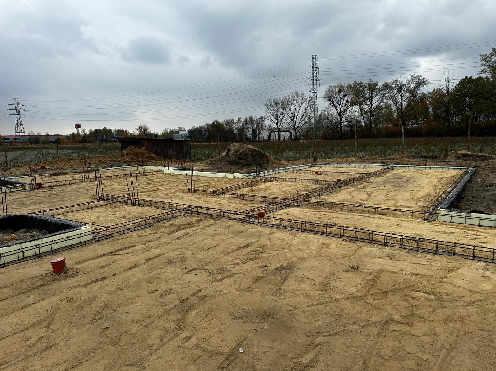 Zbrojenie fundamentów domu jednorodzinnego, widoczne stalowe pręty tworzące szkielet, szalunki z białego styropianu, przygotowany teren pod budowę w pochmurny dzień.