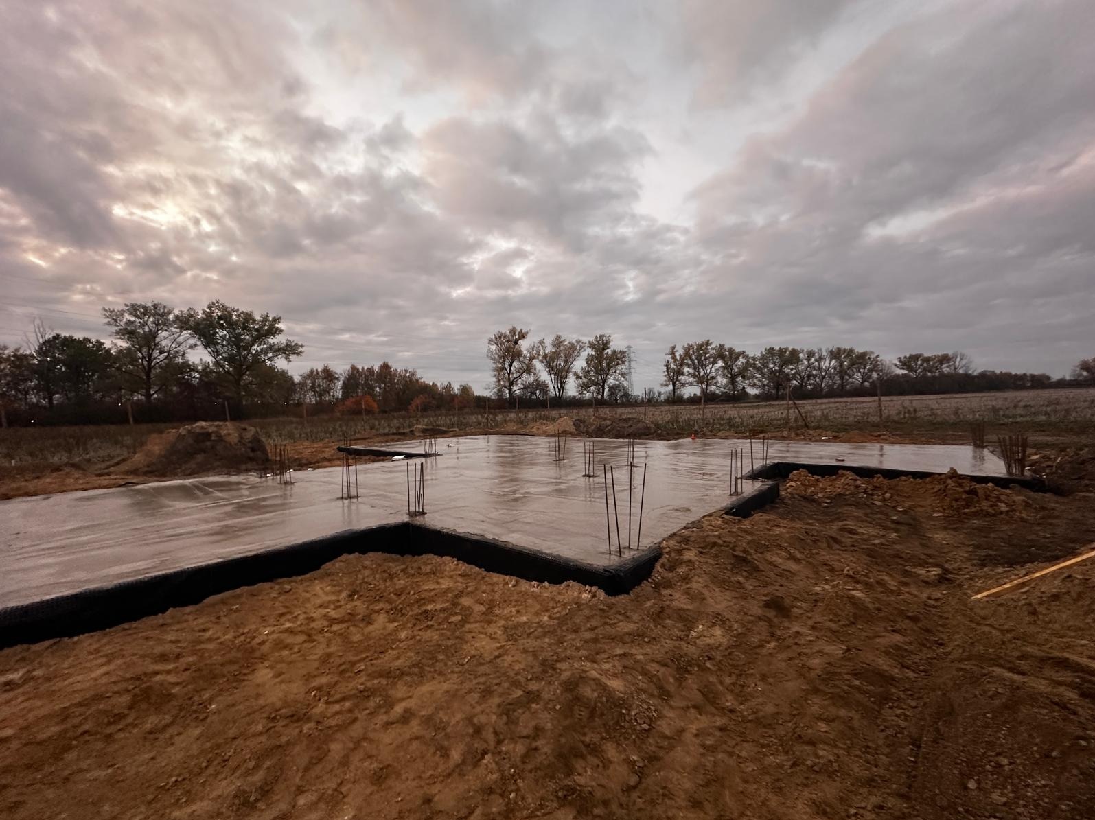 Wylany fundament z wystającymi prętami zbrojeniowymi, zabezpieczony folią, otoczony ziemią, widok na pole i drzewa pod pochmurnym niebem.