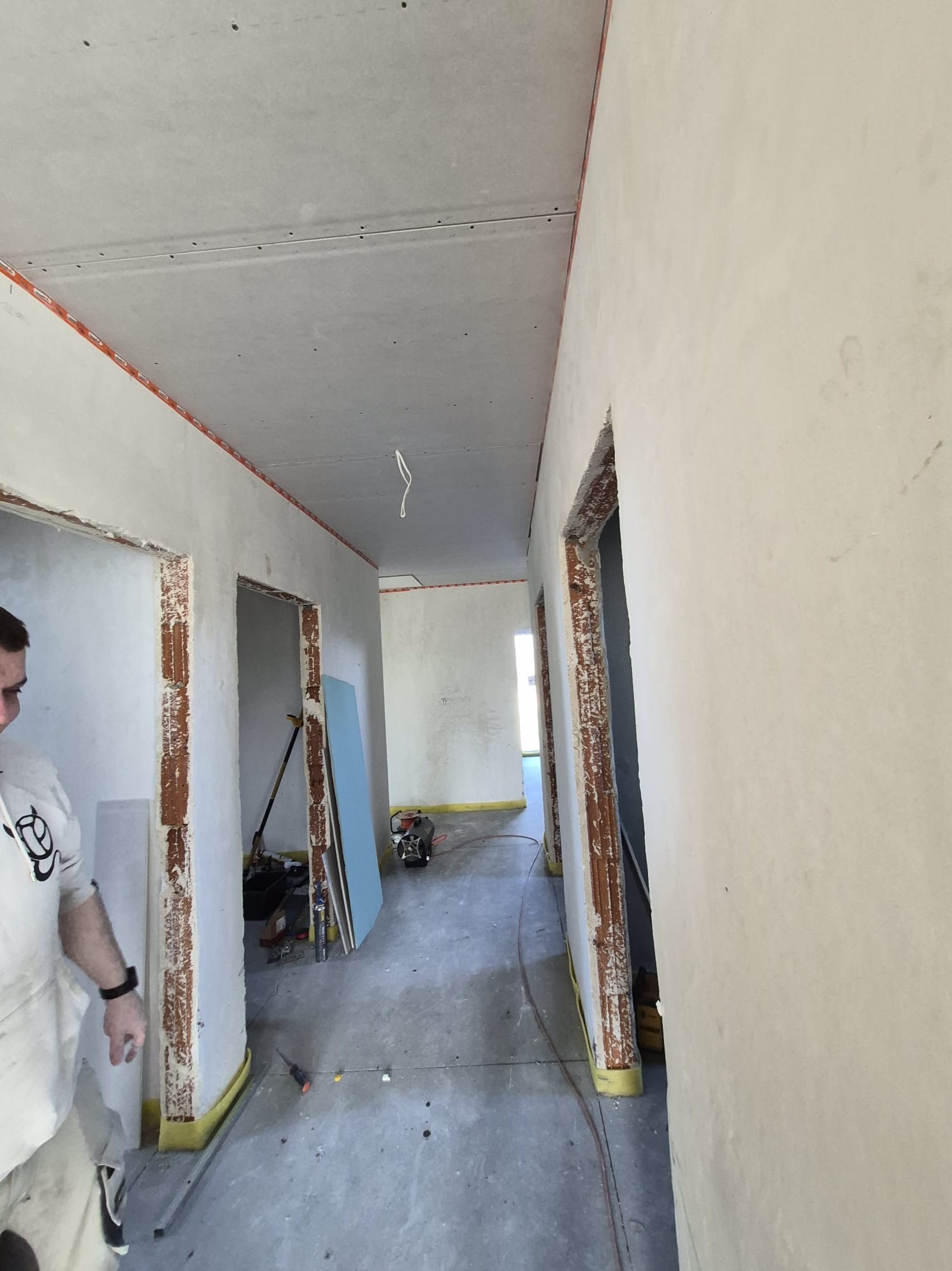 Wnętrze w trakcie prac wykończeniowych: surowe ściany, zamontowane płyty gipsowe na suficie, widoczne otwory drzwiowe i narzędzia budowlane. Etap przygotowania do malowania.