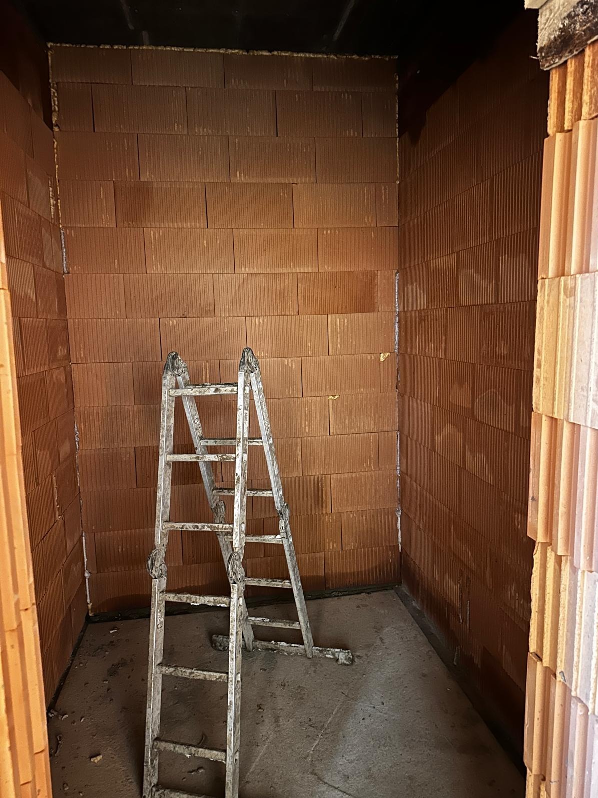 Wnętrze w trakcie budowy z cegły ceramicznej, widoczne ściany i drabina. Szare fugi między cegłami. Surowy stan, przygotowanie do dalszych prac wykończeniowych.