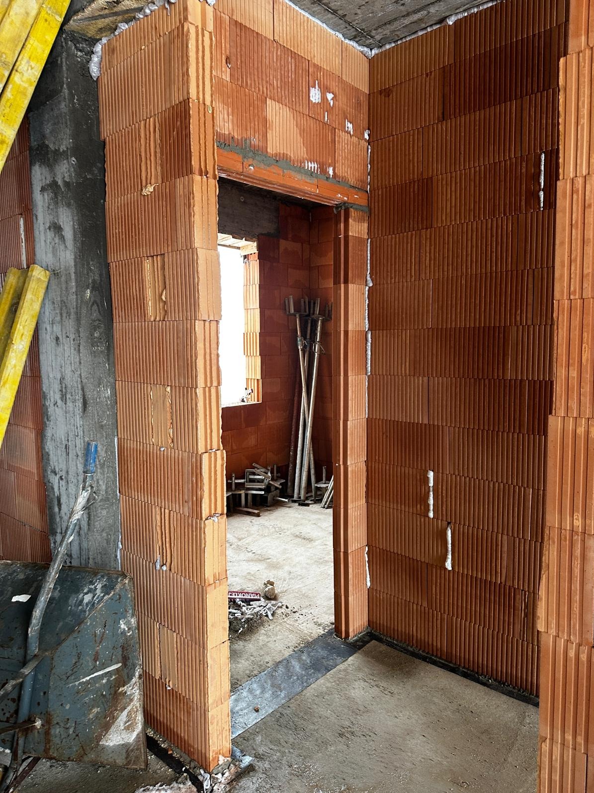 Wznoszenie ścian z pustaków ceramicznych w surowym wnętrzu. Widoczne narzędzia i materiały budowlane. Etap budowy domu z charakterystyczną fakturą cegieł.