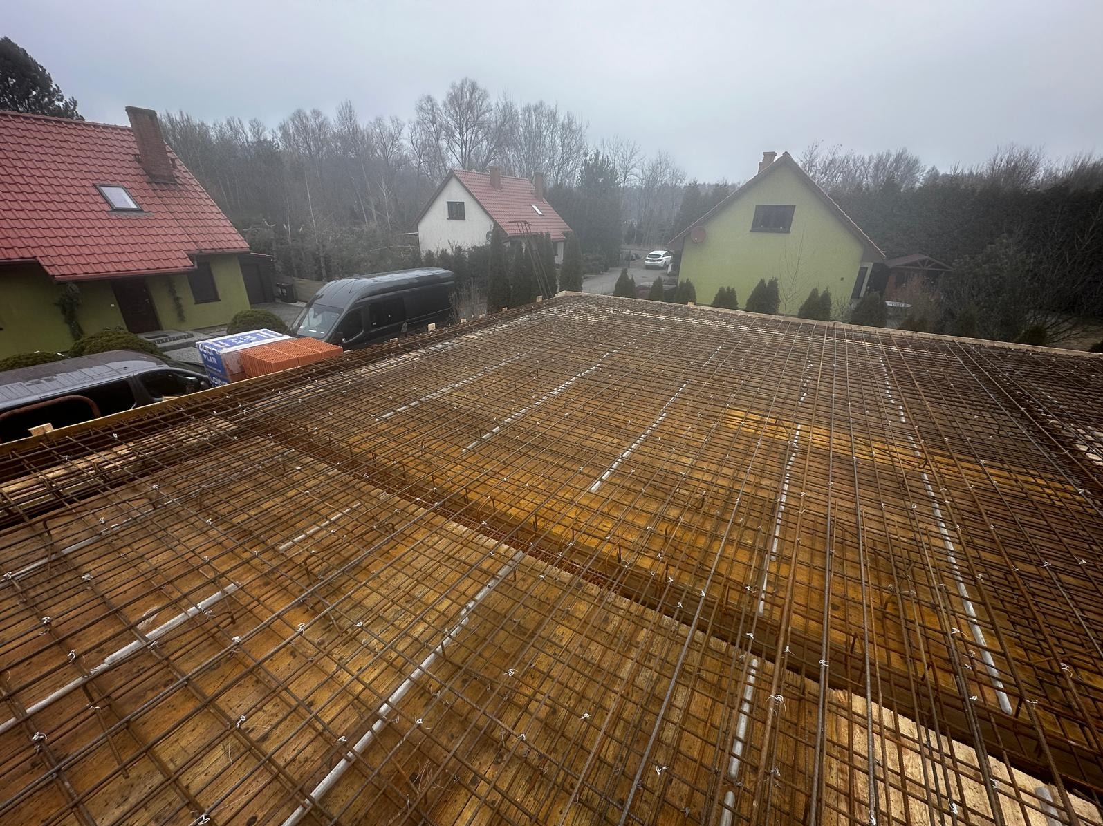 Zbrojenie stropu domu jednorodzinnego z widocznymi elementami szalunku i fragmentami okolicznych budynków w tle, wykonane w pochmurny dzień.