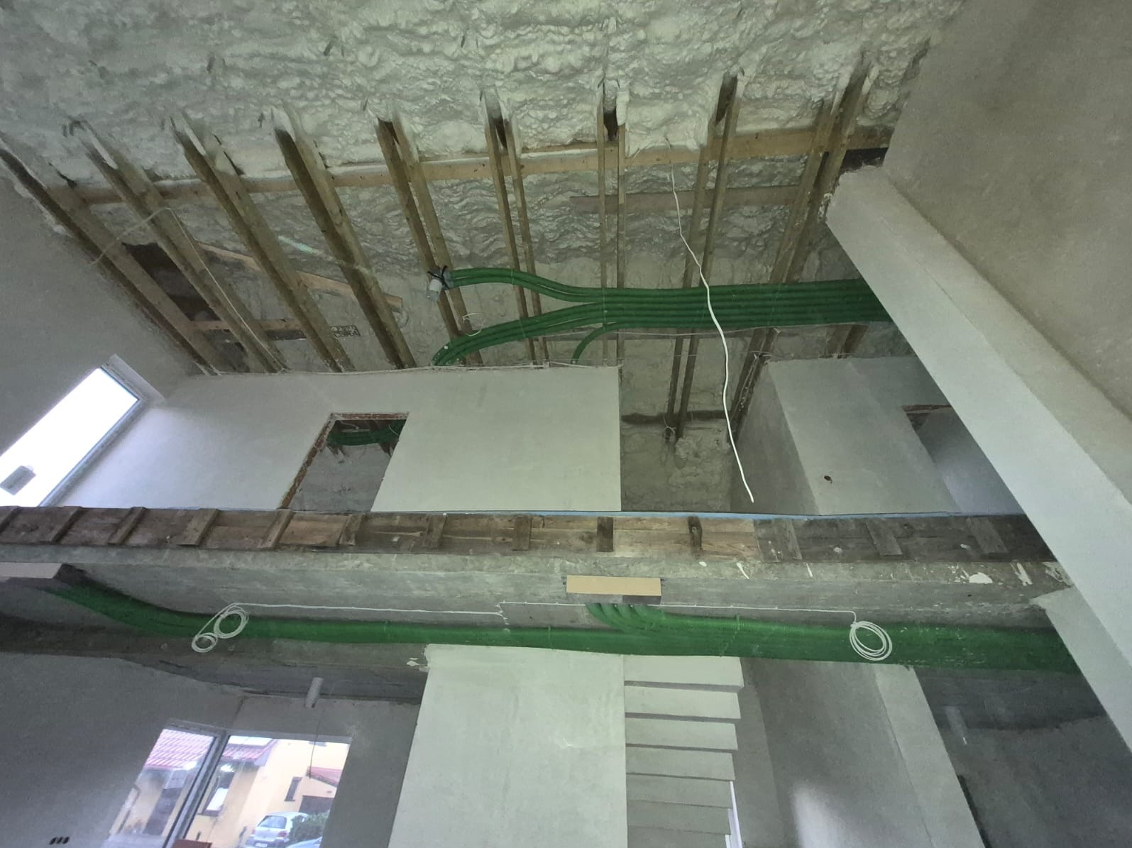 Wnętrze domu w trakcie prac wykończeniowych: ocieplony strop pianą, widoczne drewniane belki, instalacje w zielonych rurach. Surowy stan ścian, okno dachowe.