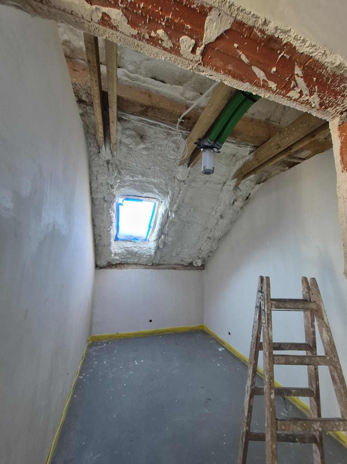 Poddasze w trakcie izolacji pianą PUR, widoczne drewniane belki, okno dachowe zabezpieczone folią, instalacja wentylacyjna i drabina. Żółta taśma na podłodze.