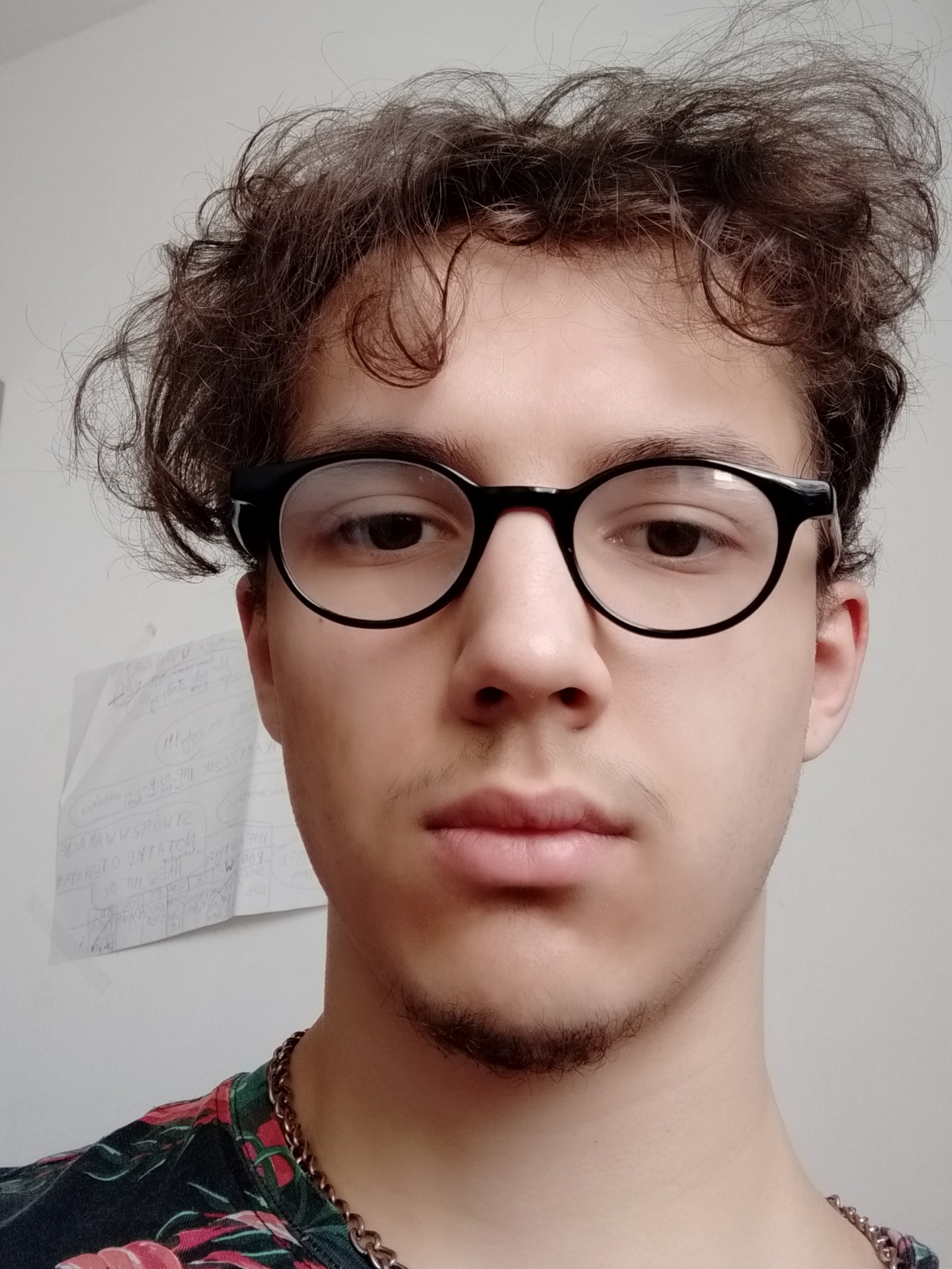 Portret młodego mężczyzny w okularach z krótką brodą, na tle białej ściany z notatkami i w kwiecistej koszuli z łańcuchem na szyi.