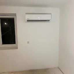 Biały klimatyzator marki Kaisai zamontowany na ścianie w nowo wyremontowanym pokoju z drewnianą podłogą, obok okna.