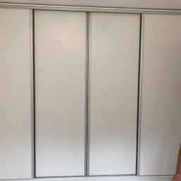 Arkadiusz Żołnowski - Czterodrzwiowa szafa wnękowa z przesuwnymi, białymi drzwiami z aluminiowymi ramami, widoczny fragment ściany w surowym stanie po lewej stronie.