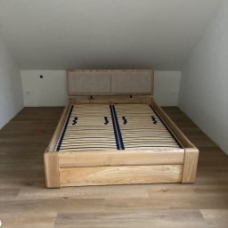 Arkadiusz Żołnowski - Drewniane łóżko z listwowym stelażem i tapicerowanym zagłówkiem, umieszczone na poddaszu z drewnianą podłogą.