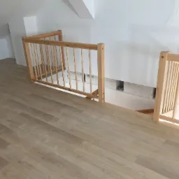 Drewniana balustrada schodów na poddaszu z jasną podłogą panelową, widoczne otwory w ścianie pod schodami.