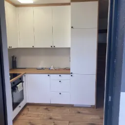 Nowoczesna kuchnia z białymi frontami i drewnianym blatem, widoczna z perspektywy wejścia do pomieszczenia, z drewnianą podłogą i czarnymi uchwytami.