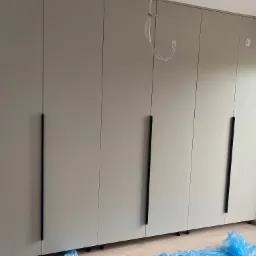 Duża szafa na wymiar z beżowymi frontami i długimi, czarnymi uchwytami, stojąca na drewnianej podłodze przy oknie. Widoczne zabezpieczenia podłogi folią.