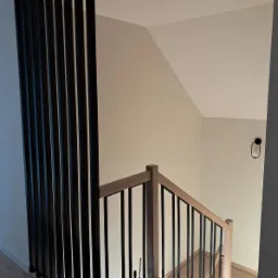 Nowoczesne schody z jasnego drewna i czarnymi, pionowymi balustradami, widziane z góry. Minimalistyczny design, neutralna kolorystyka ścian i delikatne oświetlenie.