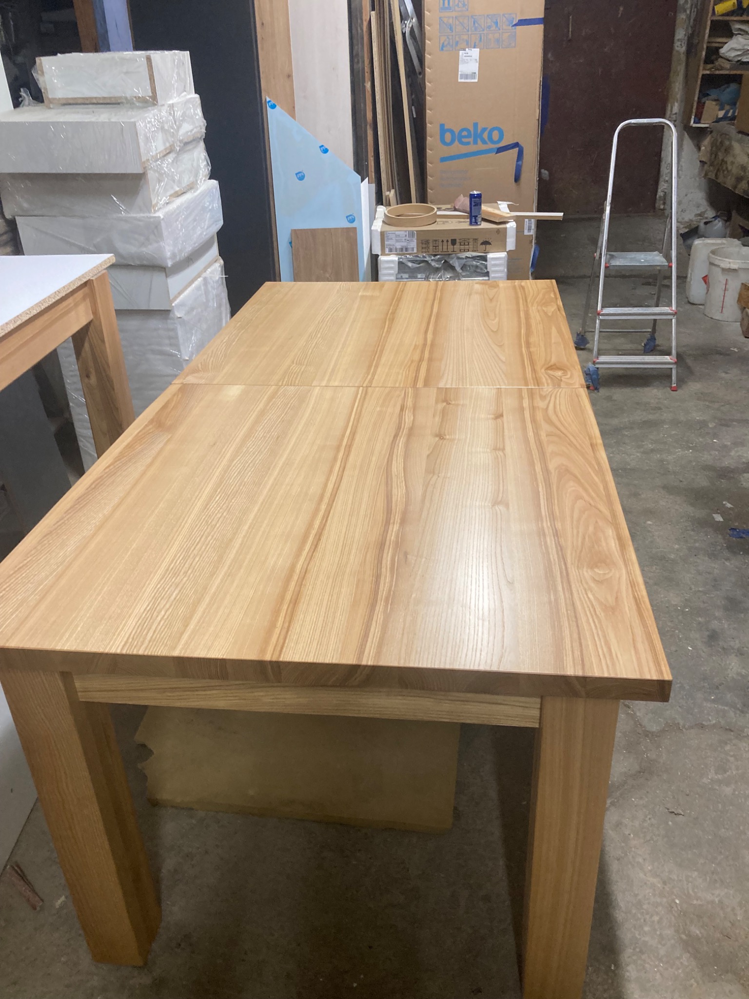 Rozkładany stół z jasnego drewna o widocznej strukturze słojów, sfotografowany w warsztacie, gdzie w tle widoczne są materiały budowlane i drabina.