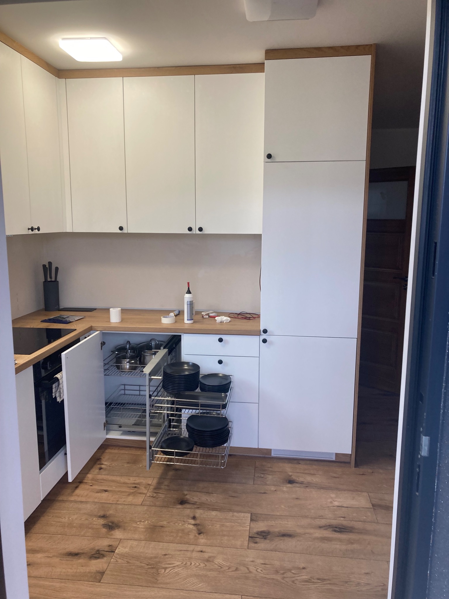 Nowoczesna biała kuchnia z drewnianymi akcentami, otwartą szafką z wysuwanymi półkami na naczynia i garnki, blatem roboczym z akcesoriami kuchennymi oraz podłogą imitującą drewno.