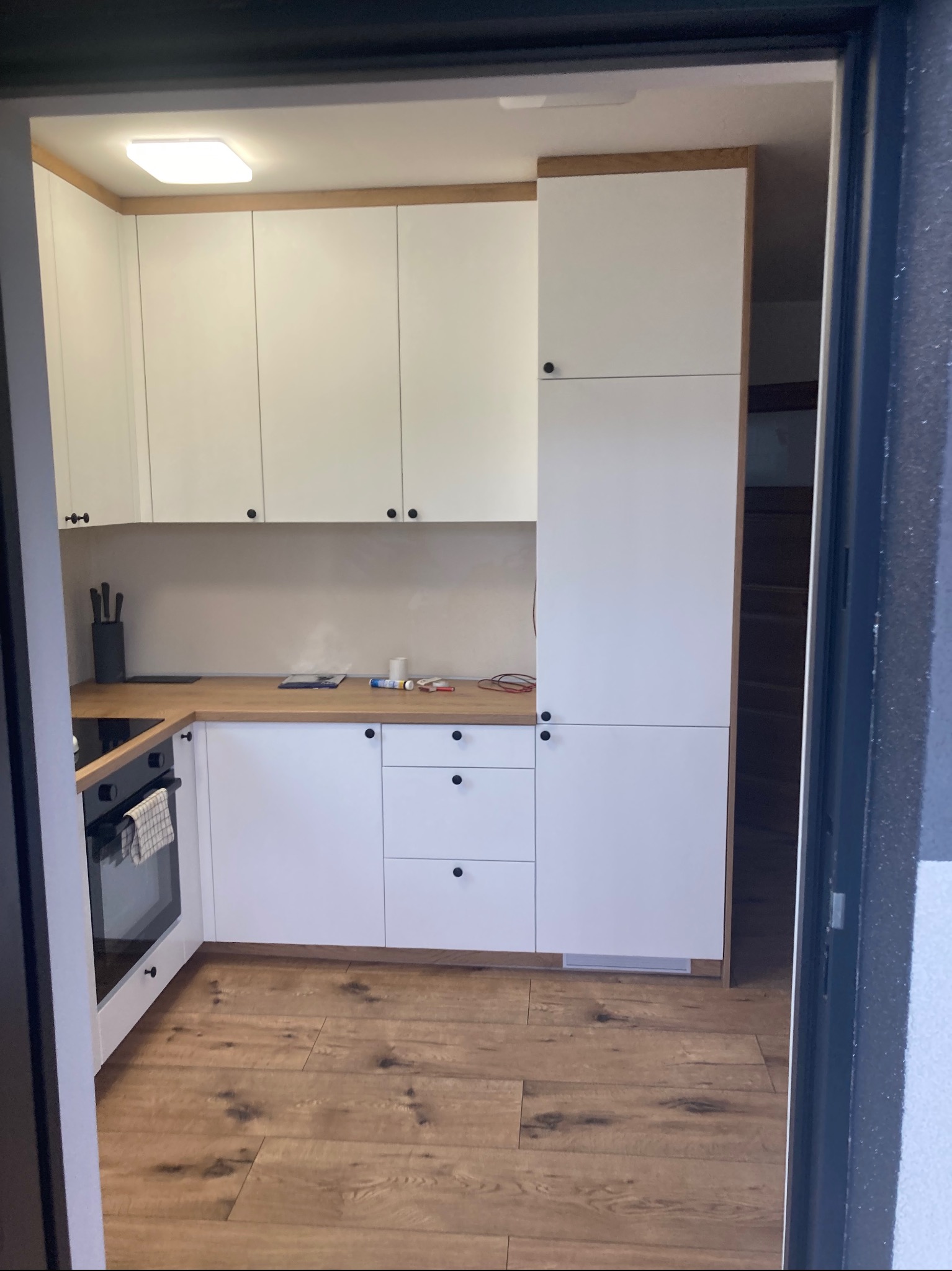 Nowoczesna kuchnia z białymi frontami i drewnianym blatem, widoczna z perspektywy wejścia do pomieszczenia, z drewnianą podłogą i czarnymi uchwytami.