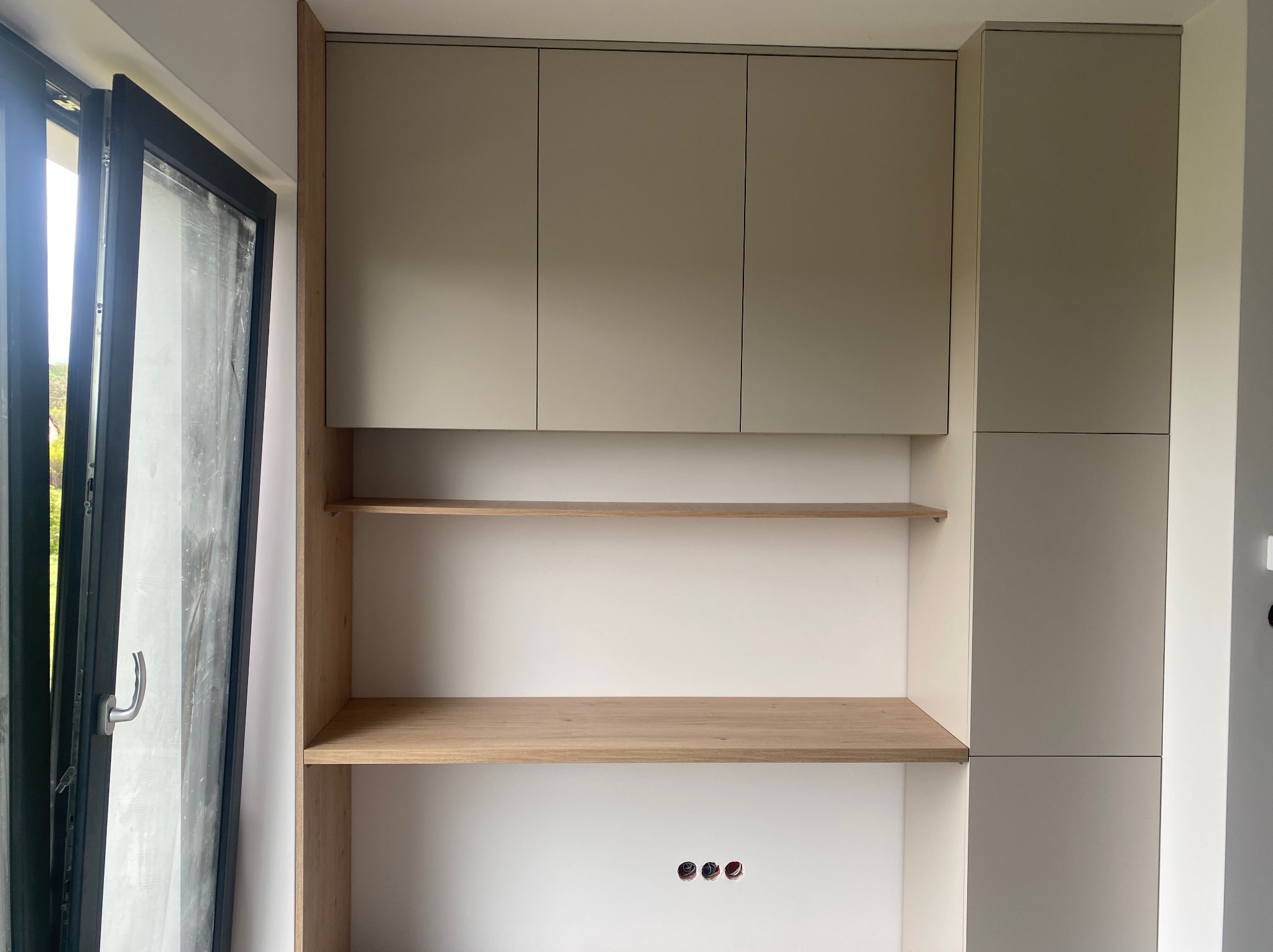 Nowoczesna szafa wnękowa z drewnianymi półkami, jasne fronty szafek, minimalistyczny design, widoczne gniazdka elektryczne, fragment okna balkonowego po lewej stronie.